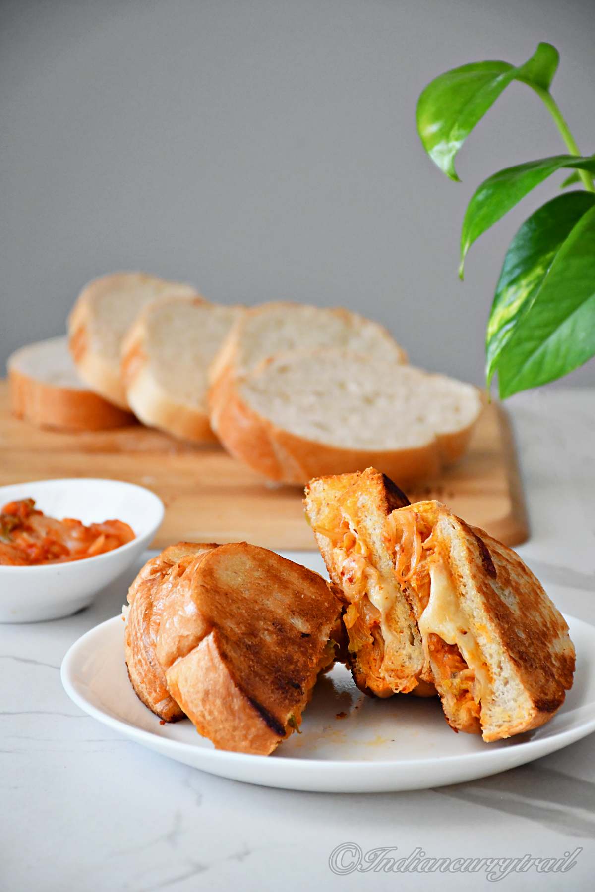 two slices of kimichi grilled cheese kept on the plate and a bowl of kimichi and bread slices kept behind
