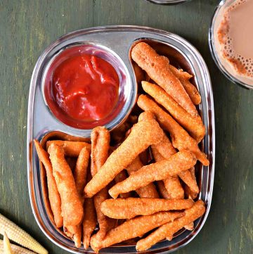 top view of baby corn golden fry, arranged on a plate with ketchup on the side and a couple of tea cups and raw baby corn kept near to it