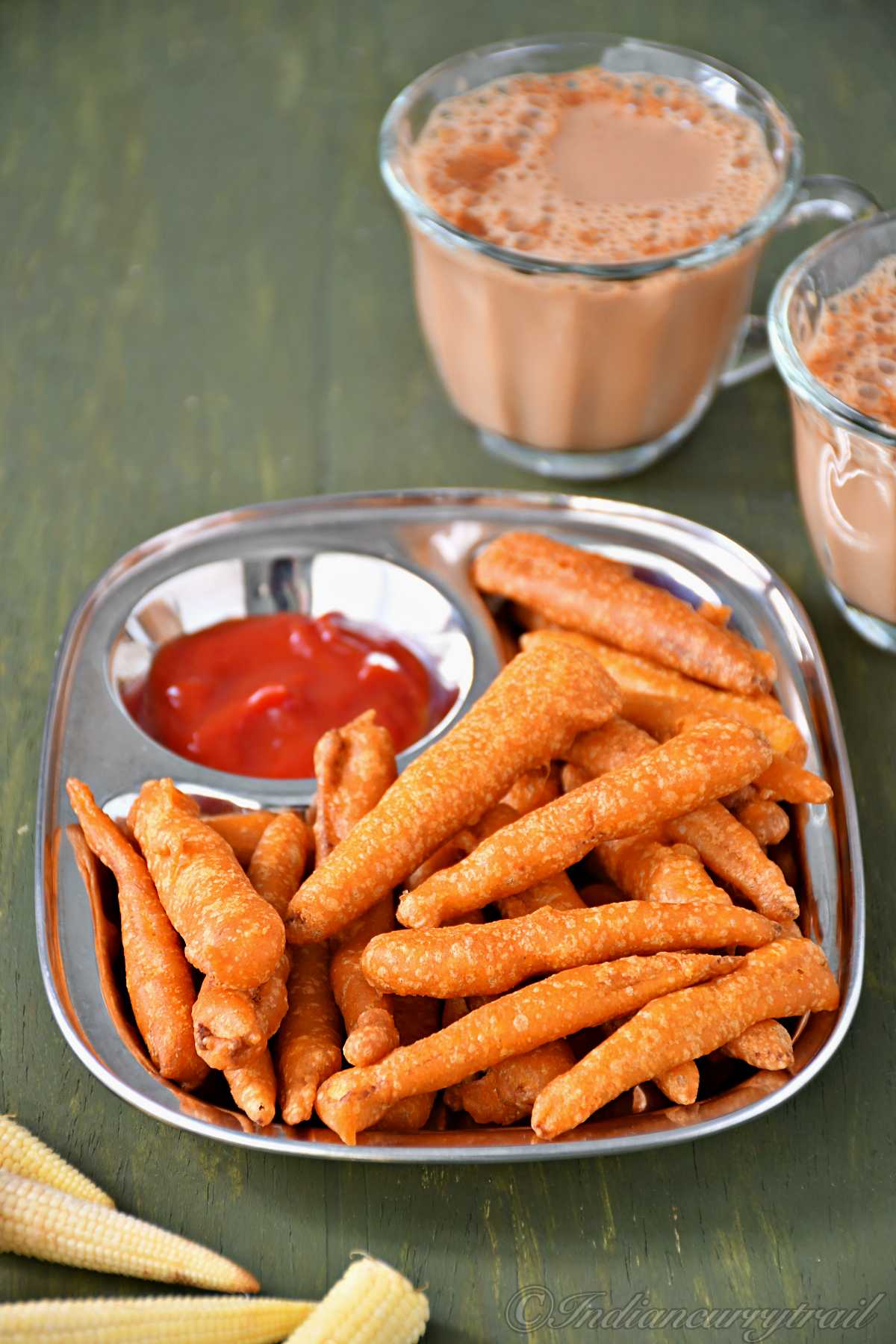 side view of baby corn fritter arranged on a plate with tomato ketchup and a couple of tea cups kept behind